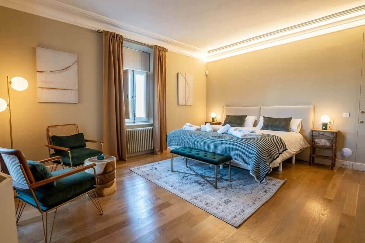 Ein Schlafzimmer mit einem großen Bett, zwei Nachttischen, einer Bank, einem Sessel, einem Heizkörper und einem Fenster mit Vorhängen.