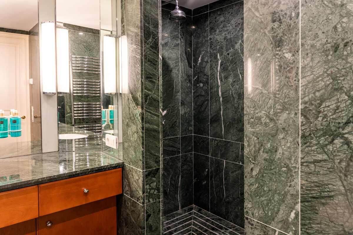 A bathroom with a glass countertop, a wooden cabinet, and a shower area with dark marble tiles.