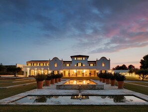 Un grand bâtiment à plusieurs étages, doté d’une façade symétrique, d’une cour centrale avec un bassin d’eau et d’une pelouse bien entretenue.