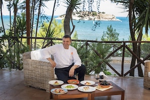 A chef seated at a table with plates of food, overlooking a coastal view.