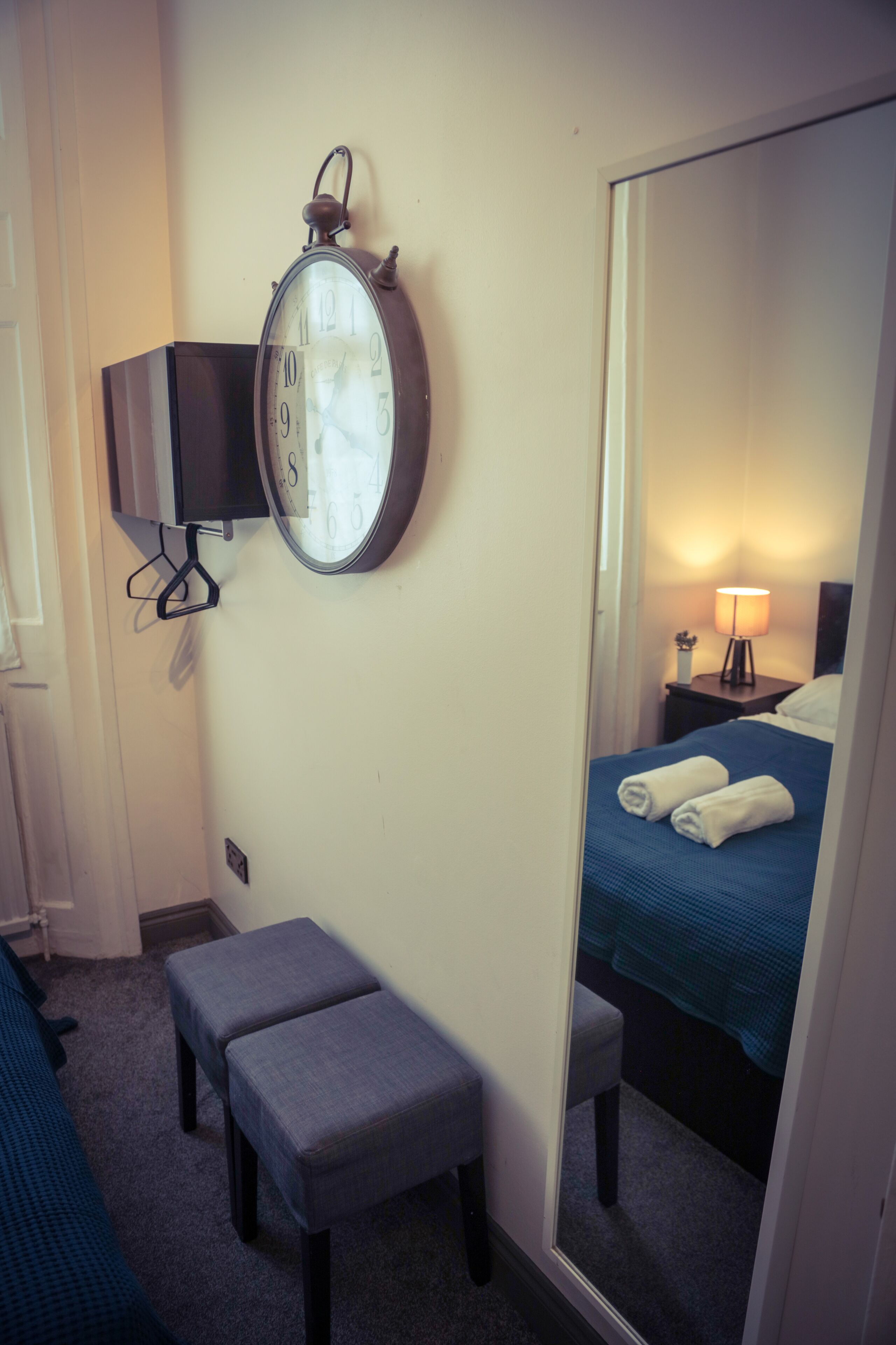A hotel room with a bed, a bedside table with a lamp, a clock on the wall, and a bench.