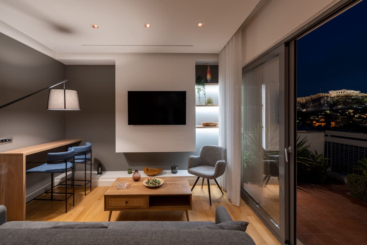 A modern living room with a flat-screen TV, a wooden coffee table, and a sofa. There are two bar stools and a chair. The room has a large window with a view of a cityscape.