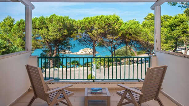 A balcony with two wooden chairs and a small table, overlooking a beach with trees and a rocky outcrop. - Gattarella Family Resort - Standard Half-Board à buffet (Vieste)