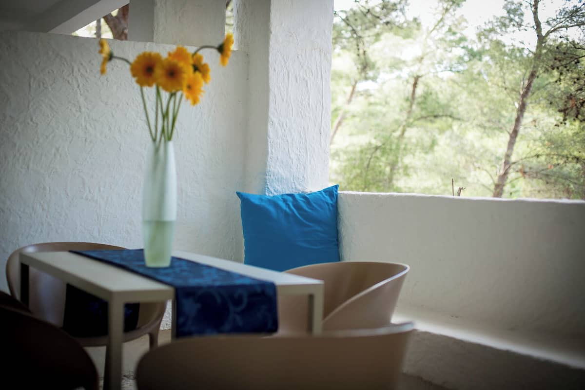 Ett rum med en vit vägg, en blå kudde, en vit vas med gula blommor, ett bord med en blå duk och ett fönster med utsikt över träd.