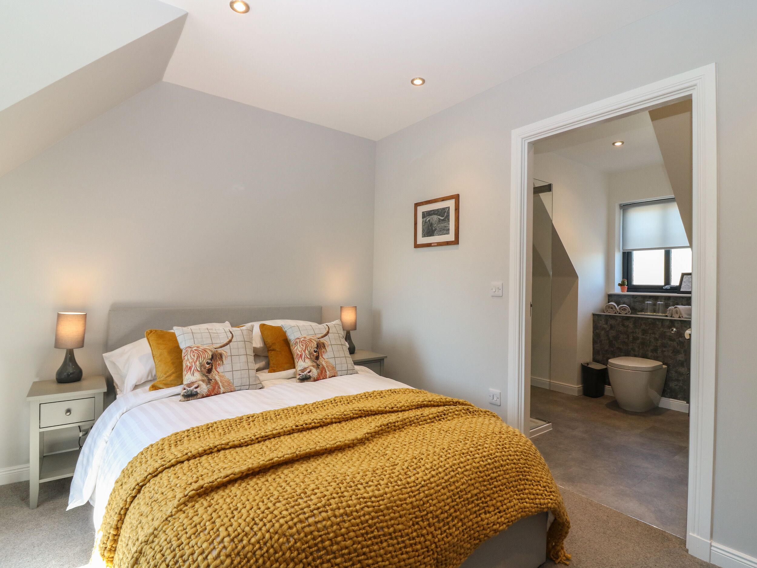 A bedroom with a bed, nightstands, a bedside lamp, a framed picture on the wall, and a bathroom visible through an open door.
