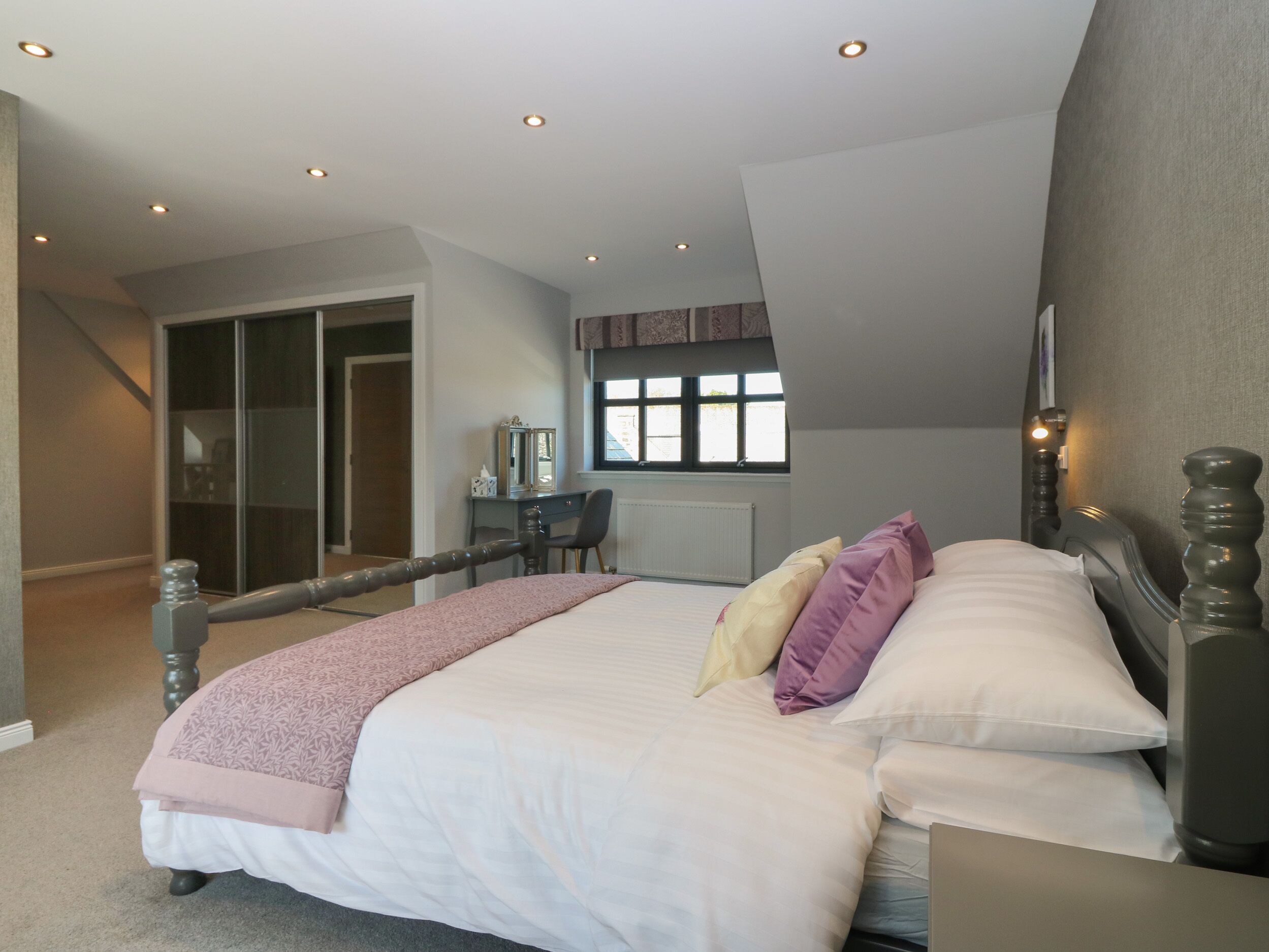 A bedroom with a large bed, a desk, and a chair. There is a window with a view of the outside.
