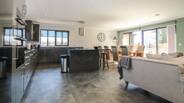 A modern kitchen with dark cabinetry, a central island, and a dining area with a sofa and chairs. - Woodend Croft (Ellon)