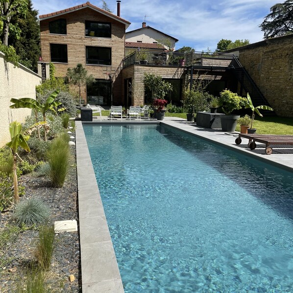 A rectangular swimming pool with clear water, surrounded by a stone wall and various plants.
