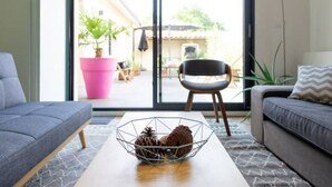 A modern living room with a sofa, a coffee table, and a chair. There is a large window with a view of a patio area. - 6-person air-conditioned gîte: terrace, garden, heated swimming pool 10 km from Lyon (Saint-Cyr-au-Mont-d'Or)