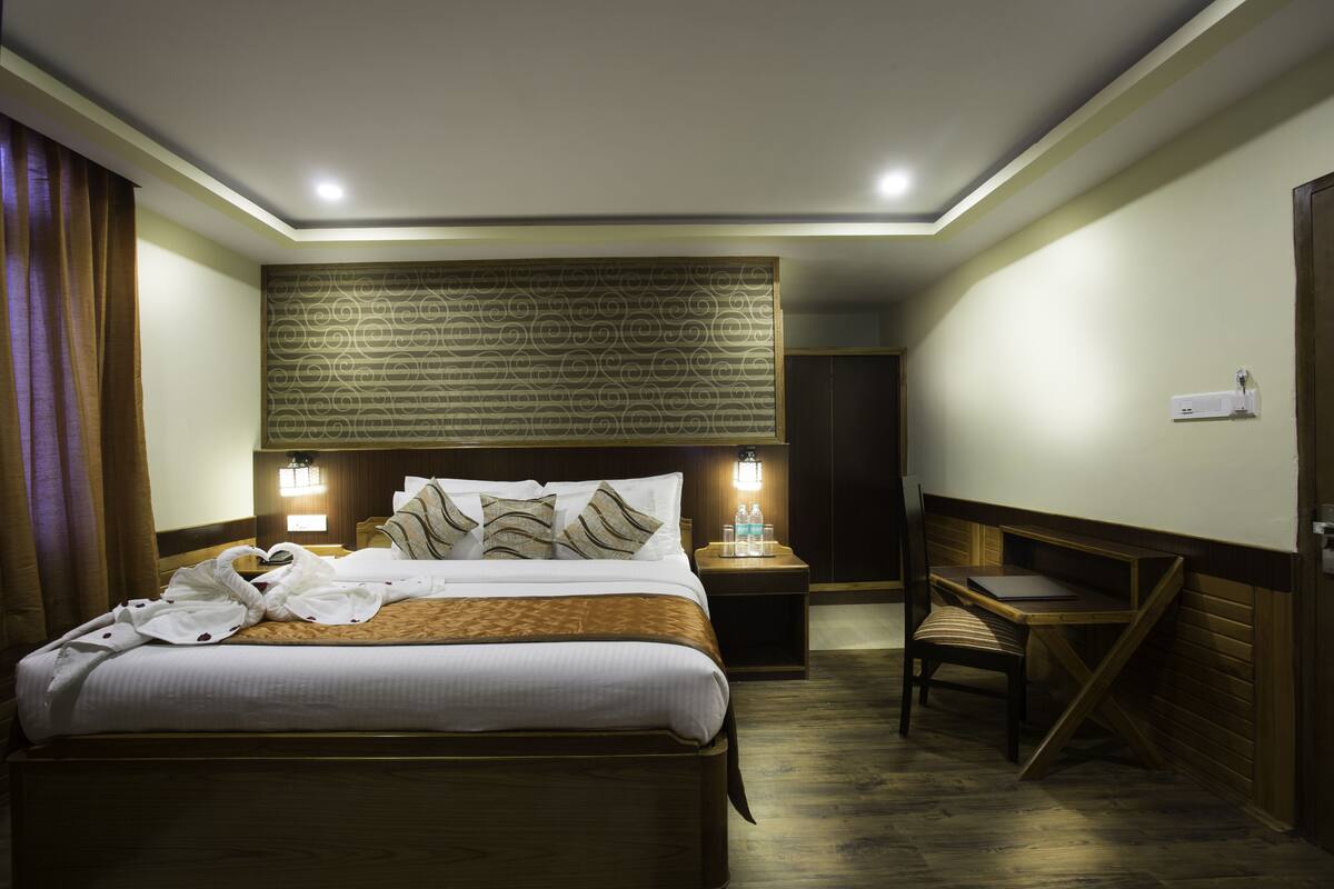 A hotel room with a neatly made bed, a desk, and a chair. There is a wall-mounted light fixture above the bed and a small table with a lamp beside the desk.