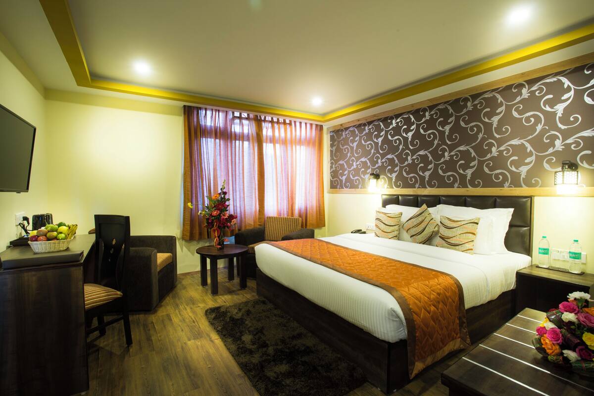 A hotel room with a bed, bedside tables, a desk with a chair, a small table with a vase of flowers, and a large window with curtains.