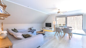 A modern living room with a sofa, coffee table, and TV. There is a dining area with chairs and a table. The room has a ceiling fan and large windows. - Fewo am Schwanenteich mit Dachterrasse (Emden)