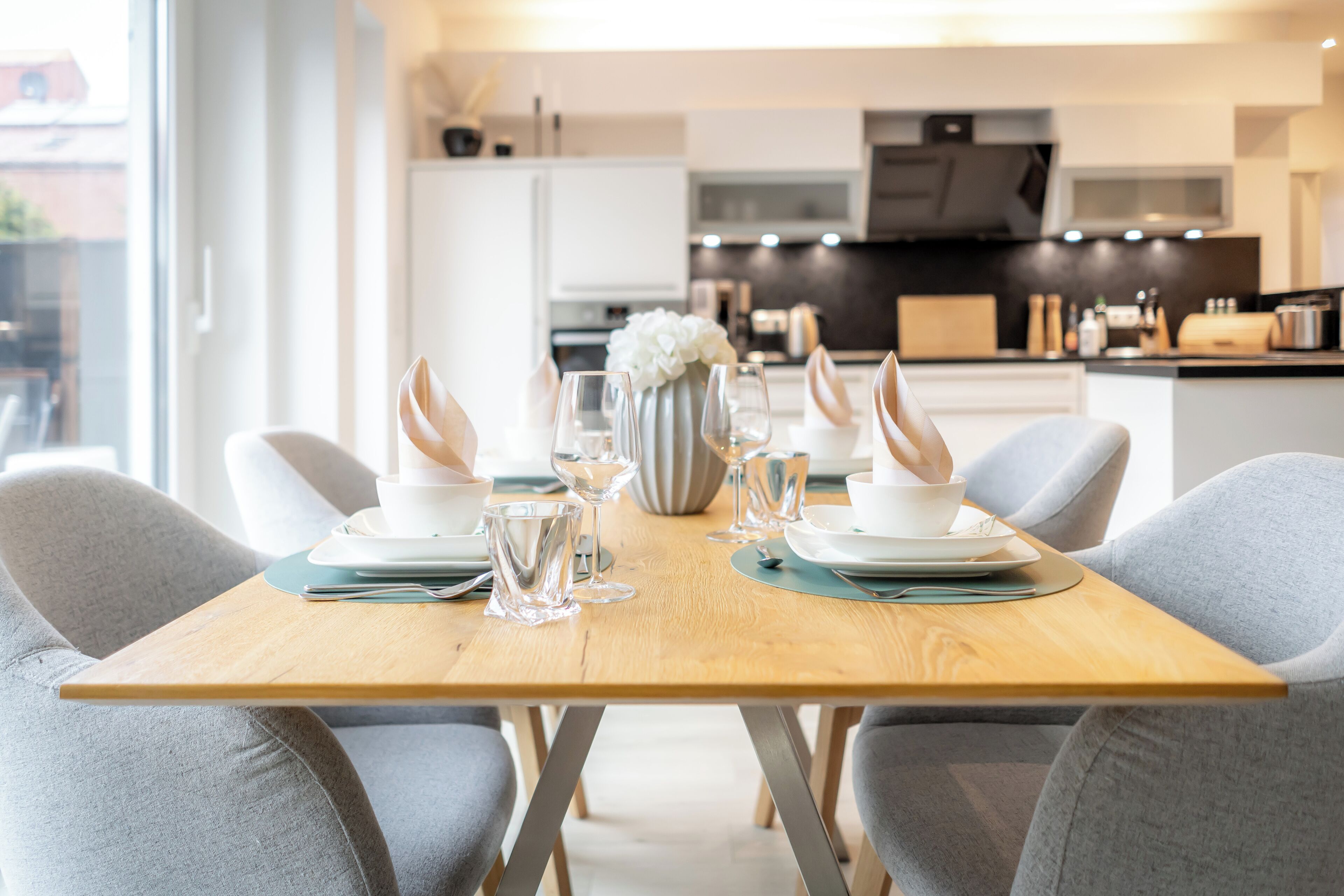 Un coin repas moderne avec une table en bois dressée pour deux personnes, agrémentée d’assiettes blanches, de verres transparents et d’un vase contenant des fleurs blanches.