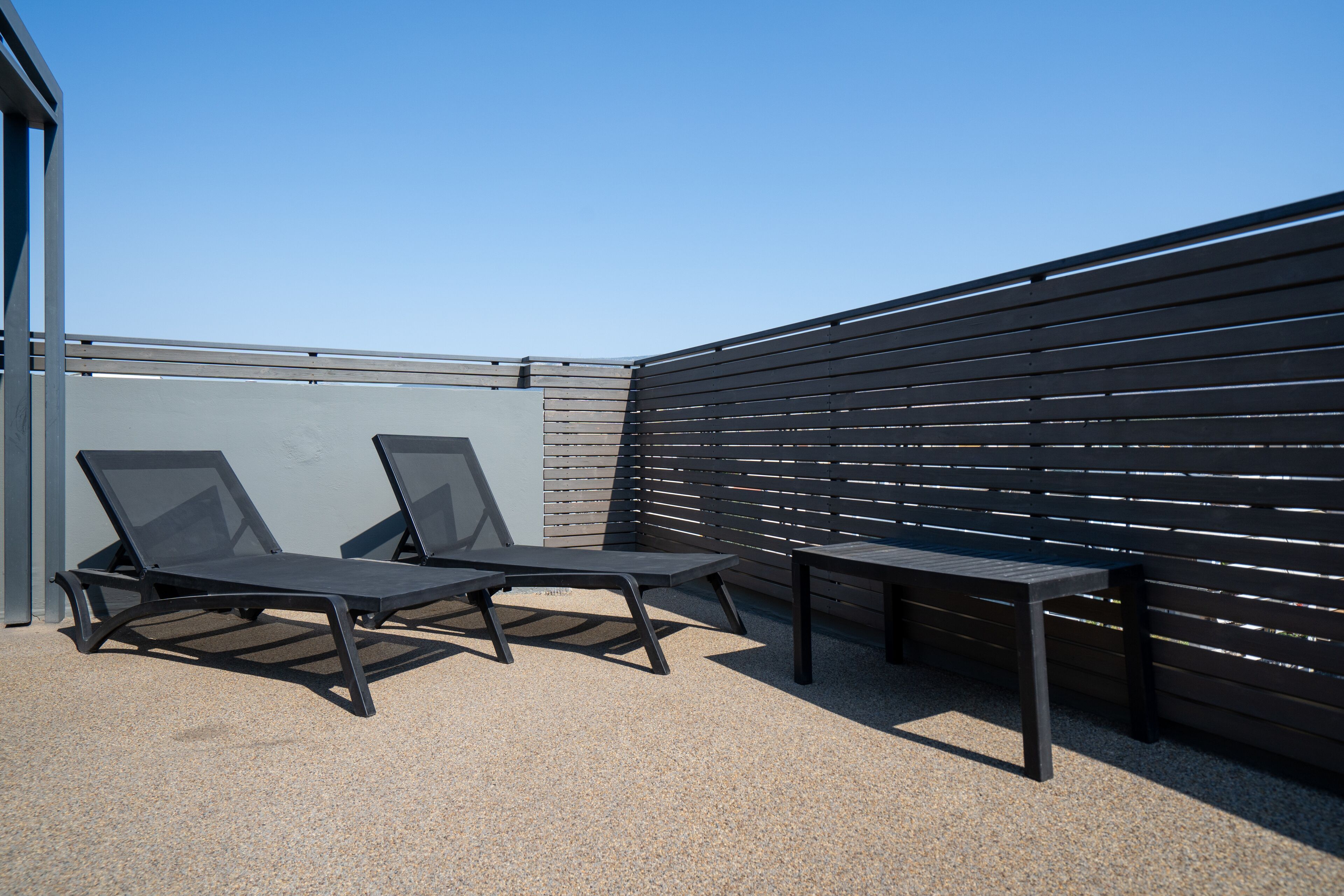 A rooftop terrace with two lounge chairs and a table, surrounded by a metal railing and a wooden fence.
