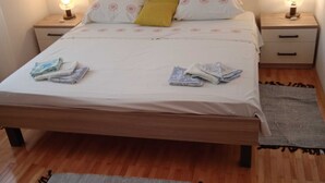 A neatly made bed with a white bedspread, two pillows, and a yellow accent pillow. There are two bedside tables with lamps, a wooden headboard, and a grey rug on the floor. - Apartments Marita - Two Bedroom Apartment (Korčula)