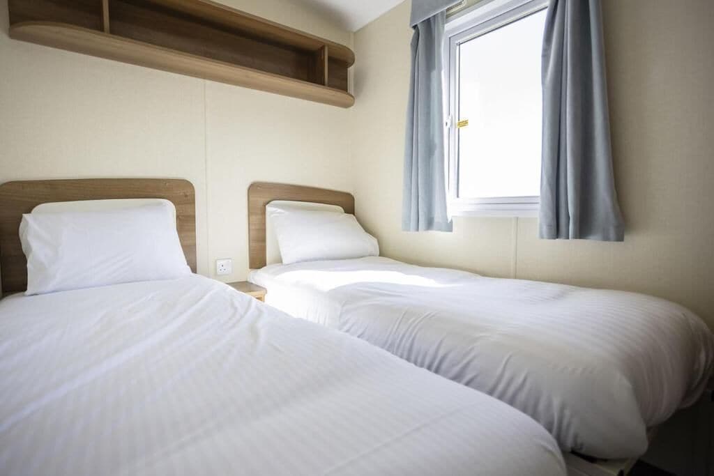 A room with two beds, a wooden shelf above, and a window with curtains.