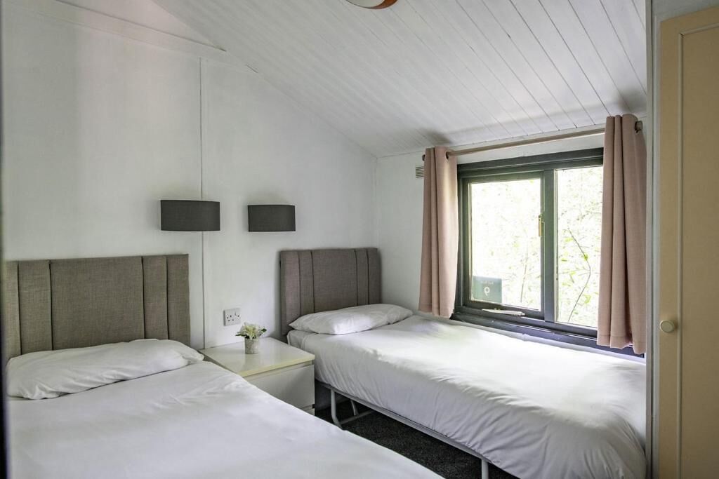 Ein Zimmer mit zwei Einzelbetten, einem kleinen Nachttisch mit Lampe, einem Fenster mit Vorhängen und einer Tür.