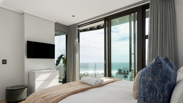 Ein modernes Hotelzimmer mit einem großen Bett, einem flachbildfernseher an der Wand und Meerblick durch raumhohe Fenster.