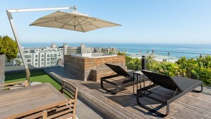 Eine Dachterrasse mit Holzboden, Liegestühlen, einem Whirlpool und Blick auf das Meer und die Stadtgebäude.