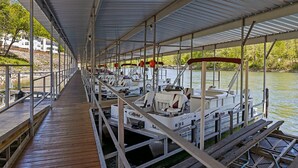 A covered dock with boats moored alongside, a wooden walkway, and a view of a river with buildings in the background.