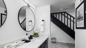 A modern bathroom with a white marble countertop, a black and white sink, a round mirror, and a black staircase with a white wall and a framed picture.