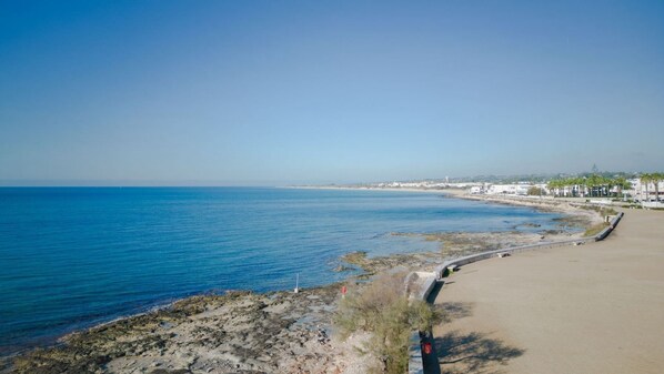 A coastal road with a beach on the right, a clear sky, and distant buildings.