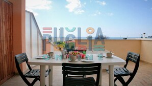 A round table with four chairs, a vase of flowers, and various dishes and glasses on a balcony overlooking the sea.