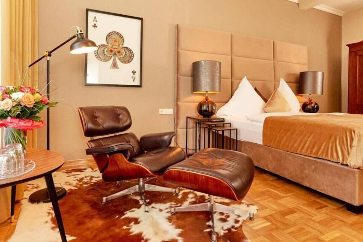 A hotel room with a bed, two chairs, a table with a vase of flowers, a lamp, and a framed playing card on the wall.