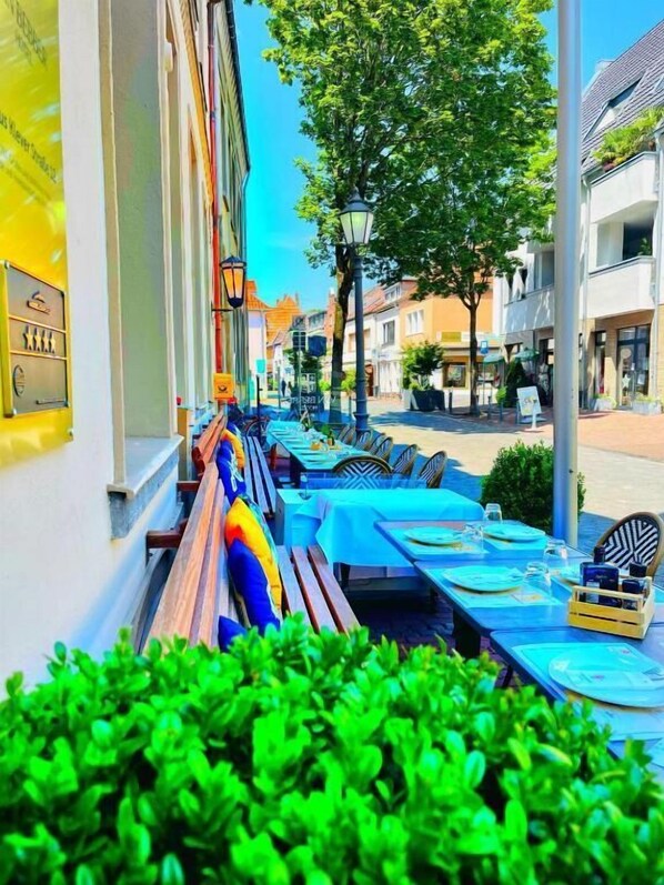 Un espacio de comedor al aire libre con mesas y bancos, vista a la calle con edificios y un cielo despejado.