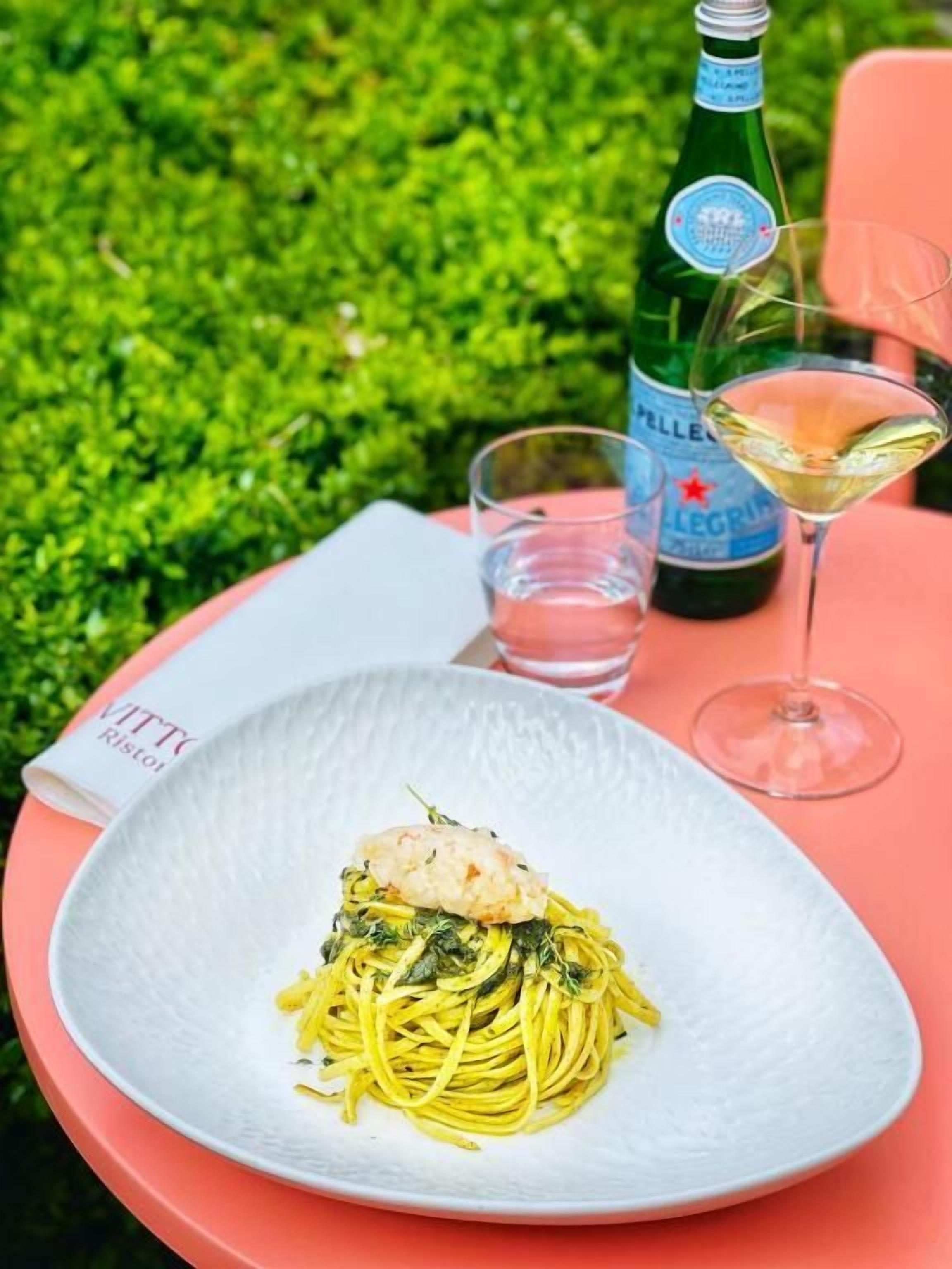 Un piatto di spaghetti con gamberetti, una bottiglia di acqua Pellegrino e un bicchiere di vino bianco su un tavolo rosa all'aperto.