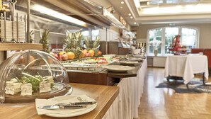 A buffet area with a variety of food items, including cheese and fruits, in a well-lit room with a wooden floor and a view of the outdoors. - Hotel Van Bebber (Xanten)
