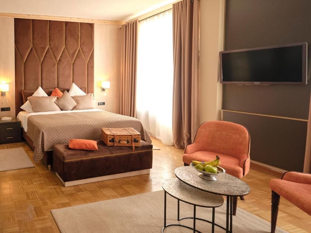A modern hotel room with a large bed, a flat-screen TV, a small round table with fruit, and a comfortable armchair.