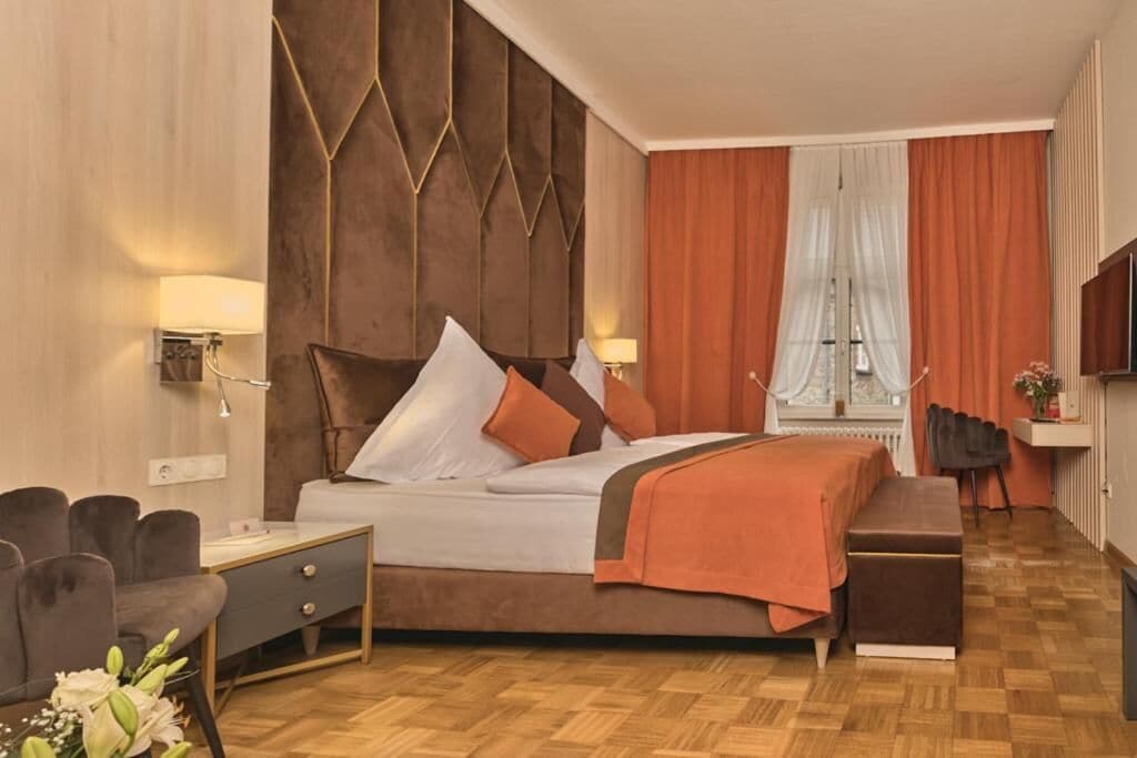 A hotel room with a bed, bedside table, lamp, chair, and a view of the city through the window.