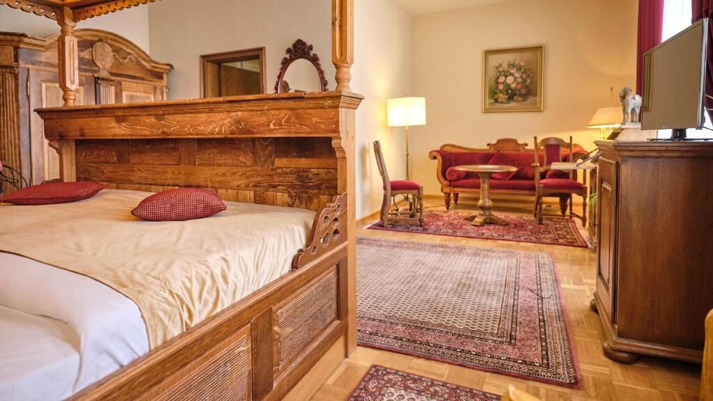 Una cama tradicional con dosel y un cabecero de madera, con un cojín rojo y blanco a cuadros y otro a juego en la cama. A la derecha, un mueble de madera, un sofá rojo y un televisor sobre un pedestal de madera.