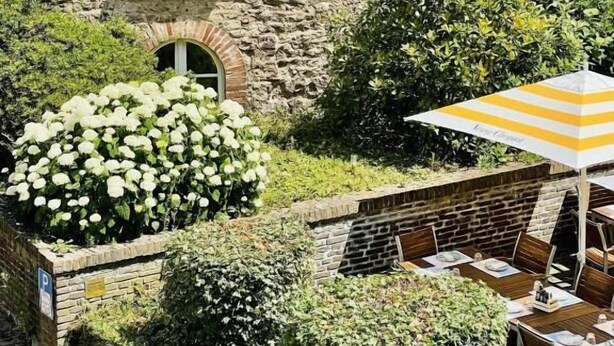 A stone building with a window, a hedge, and outdoor seating with an umbrella. - Hotel Van Bebber (Xanten)