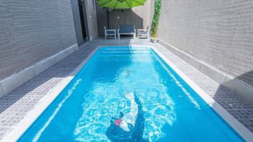 Una piscina con plataforma de baldosas, un banco y un parasol verde.