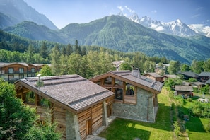 Un chalet de montagne doté de cabanes en bois et de structures en pierre, entouré d’une végétation luxuriante et de montagnes aux sommets enneigés.