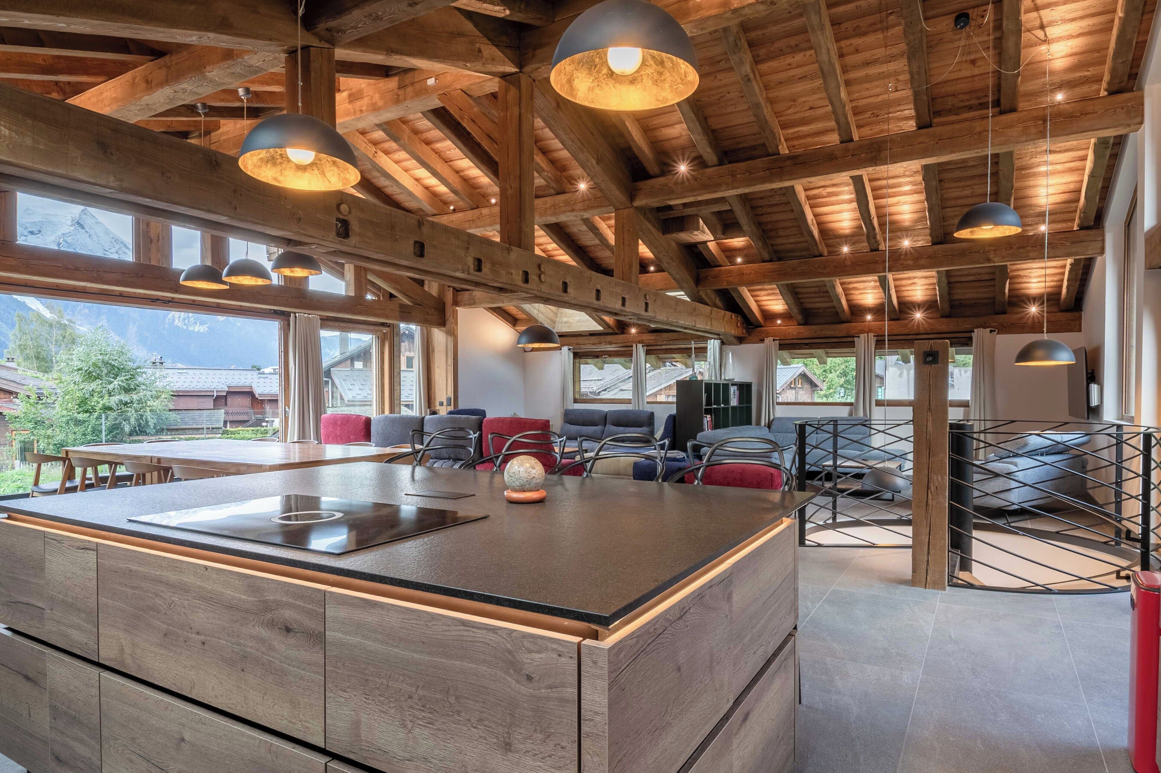 Un'ampia cucina con isola centrale, travi in legno a soffitto e lampade a sospensione, e ampie finestre che offrono vista sulla campagna circostante.