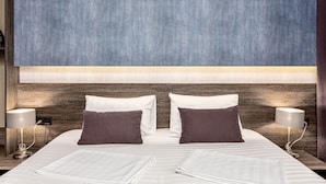 Un lit double avec du linge de lit blanc et deux oreillers foncés, de part et d’autre, deux lampes de chevet.