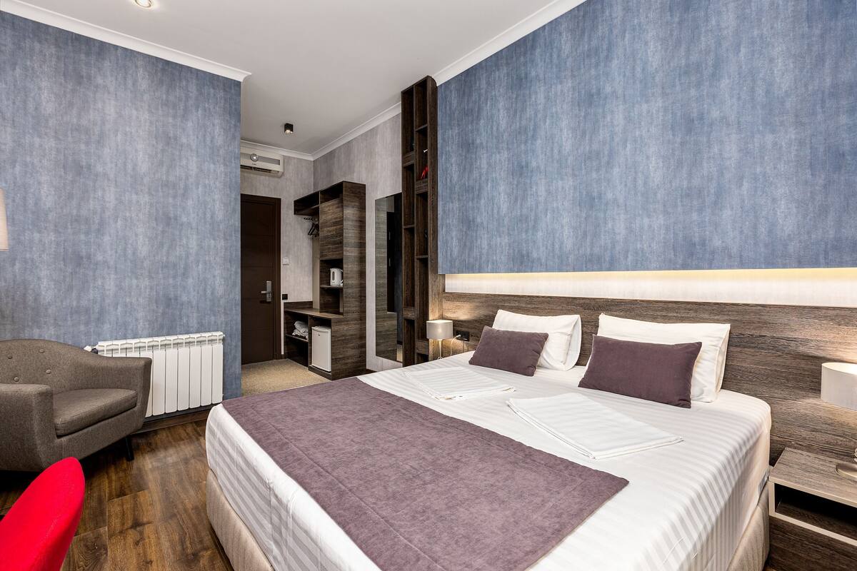 A modern hotel room with a large bed, a red chair, a wardrobe, and a built-in shelf unit.