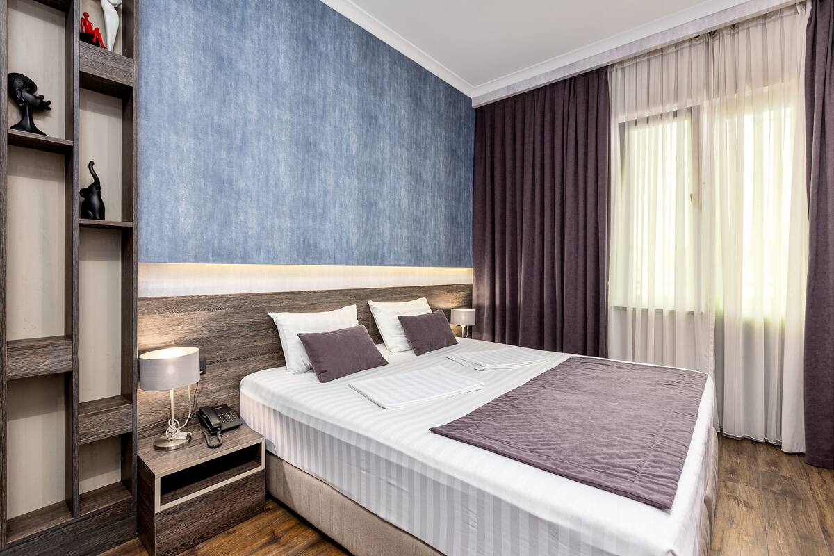A neatly made bed with white linens and a grey throw, a bedside table with a lamp and phone, and a blue accent wall.
