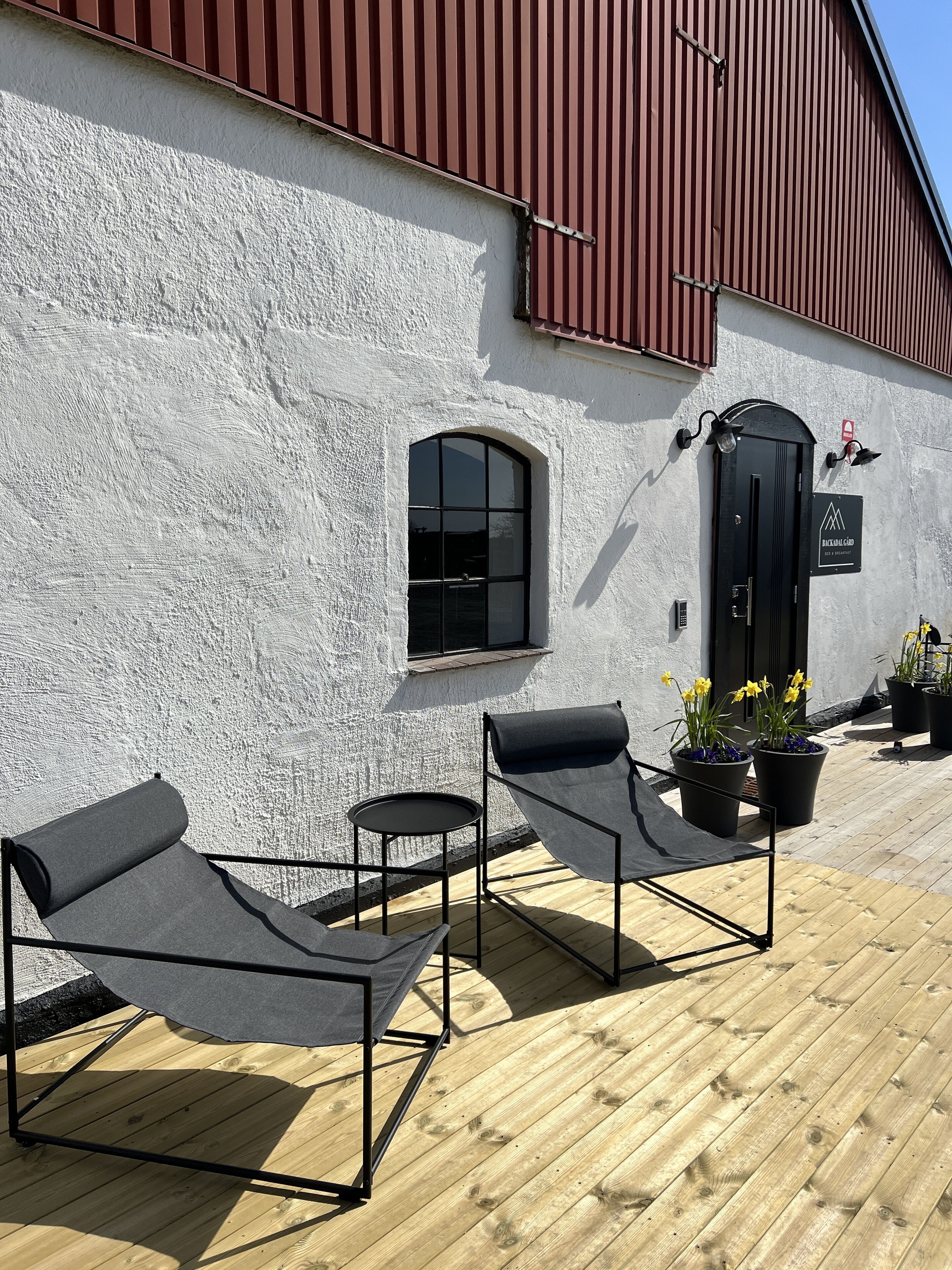 A building with a white exterior and a red roof. There are two black lounge chairs with a small table and a potted plant with yellow flowers.