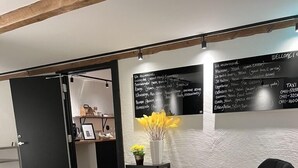 Un área de espera moderna con un sofá negro, una pequeña mesa redonda y un florero decorativo con flores amarillas. Un pizarrón con texto está montado en la pared.
