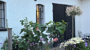 Un edificio blanco con puerta y ventanas negras, rodeado de un jardín con diversas plantas y flores.