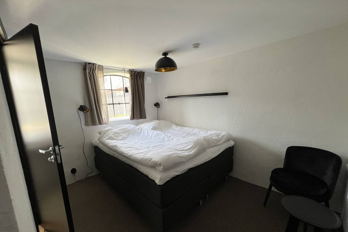A bedroom with a bed, a chair, a window with curtains, a ceiling fan, and a wall-mounted shelf.