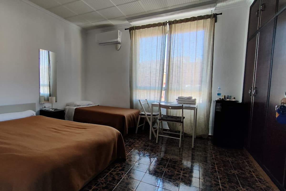A hotel room with two single beds, a small table with a chair, a desk, and a wardrobe.