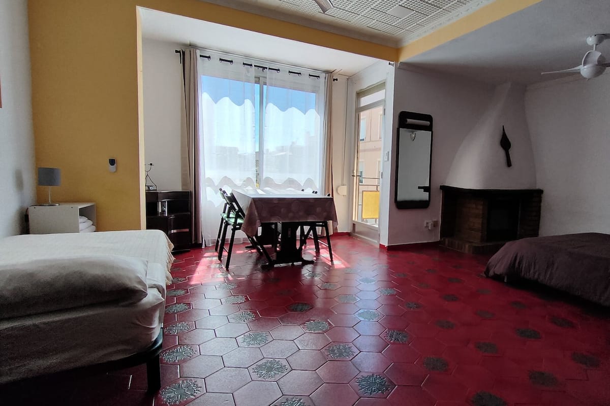 A hotel room with a red hexagonal floor, a bed on the left, a table with chairs, a fireplace, and a window with curtains.
