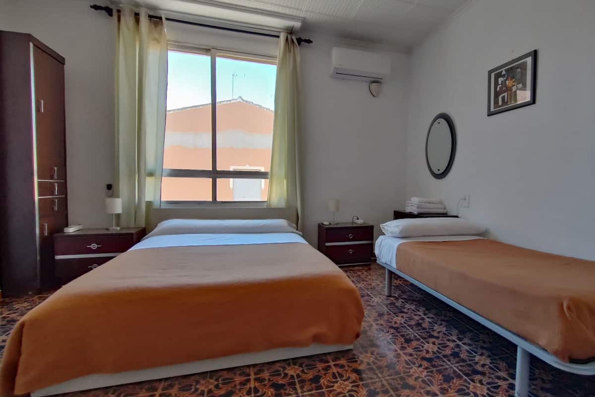 A hotel room with two beds, a wardrobe, a window with curtains, an air conditioning unit, a bedside table with a lamp, and a framed picture on the wall.