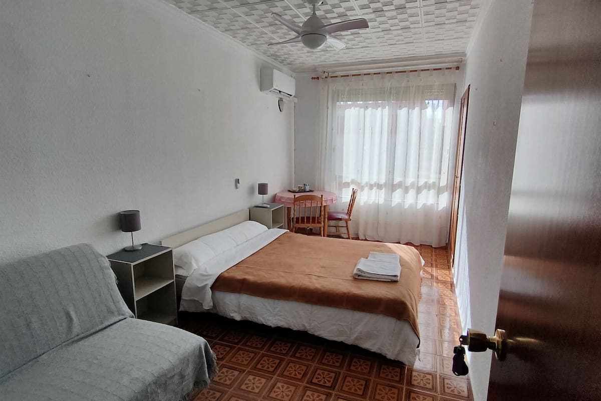 A hotel room with a bed, a chair, a small table, a desk, and a ceiling fan.