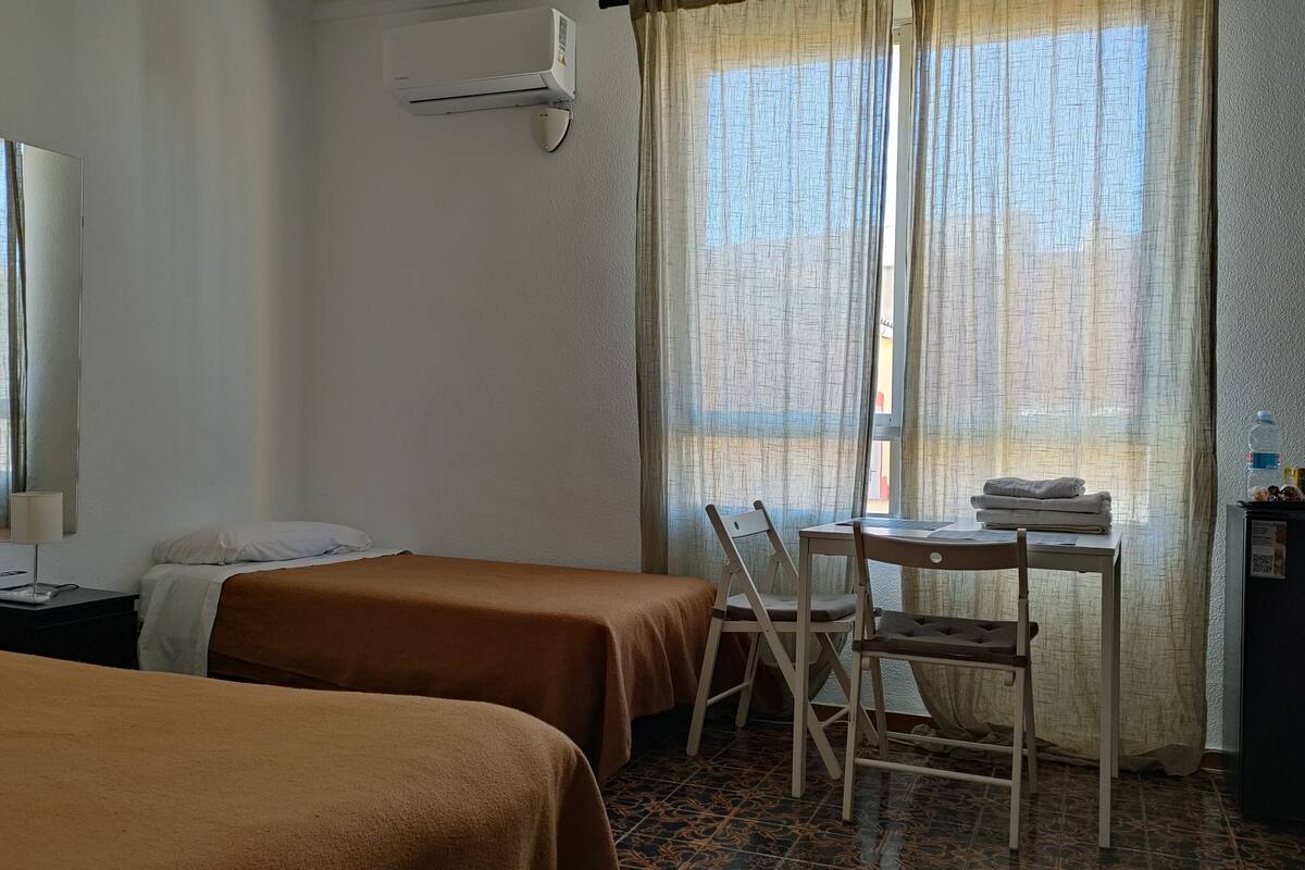 A hotel room with two beds, a desk with a chair, a mirror, and a window with curtains.